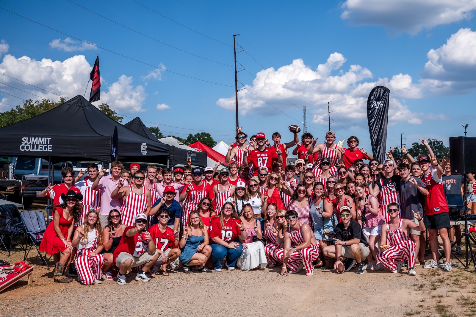 NC State tailgate group in red and white, cheering under a blue sky.