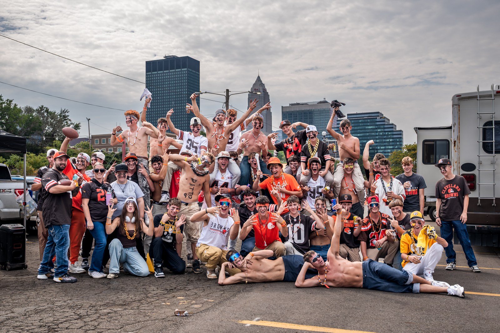 Browns tailgate group with adults at the left, fans raising their arms, and the Cleveland skyline behind them.