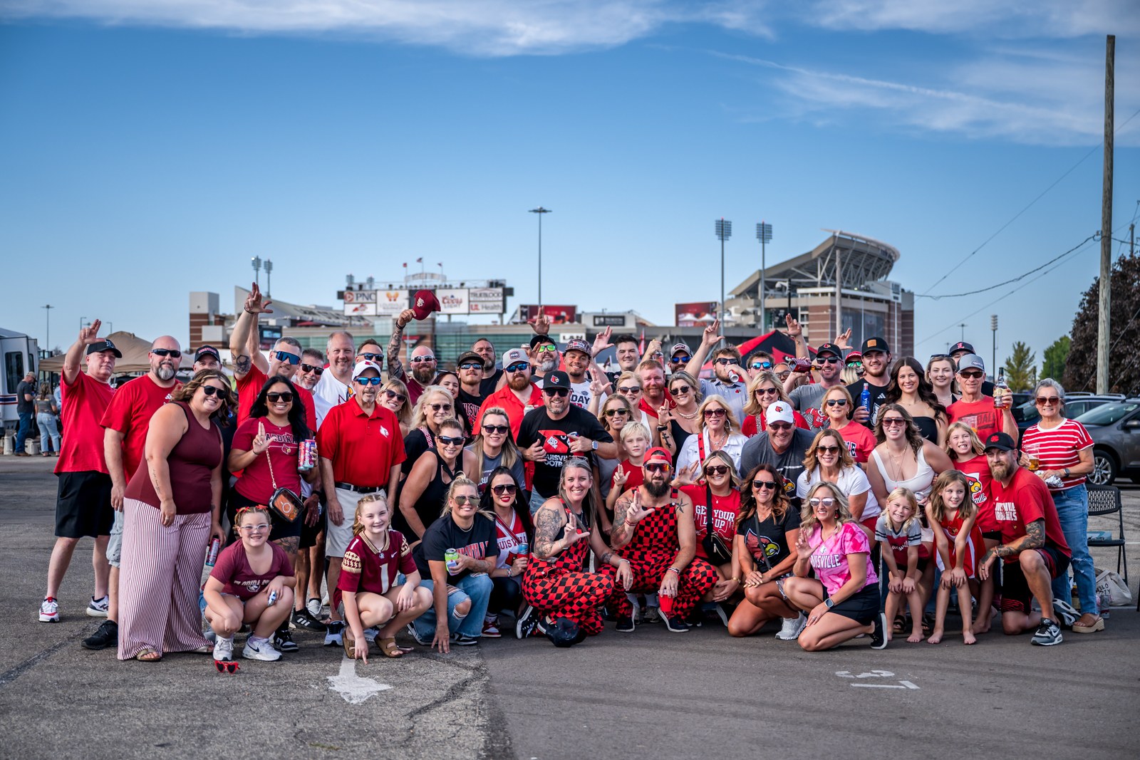 Large Louisville tailgate group in red, with a fan raising a red cap and L&N Stadium behind them.